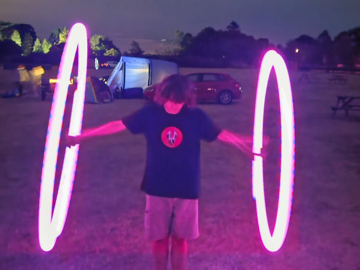 Zac spinning LED poi in a field at night, wearing an Unførgiven t-shirt, the light trails drawing two bright rings either side of him.